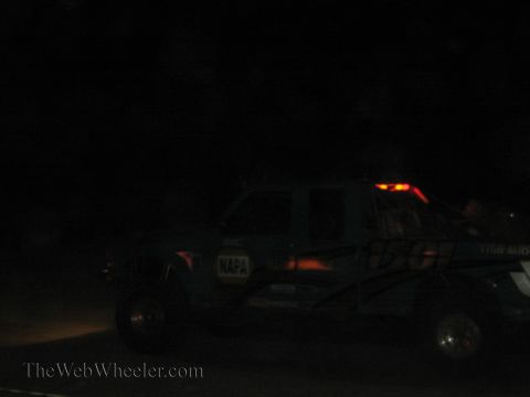 1990 Ford Bronco - lifted race pickup with a NAPA sticker and glowing roof light bar sits in near-darkness at a nighttime off-road event.