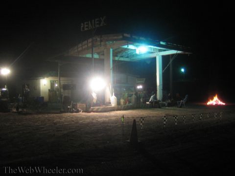 1990 Ford Bronco - Pemex gas station glows under floodlights at night beside a bonfire, lawn chairs, and a coned-off sandy lot.