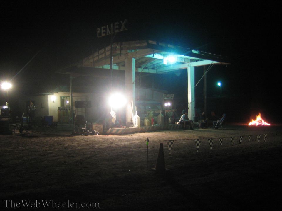 Lit Pemex fuel station at night with a bonfire and people in the desert