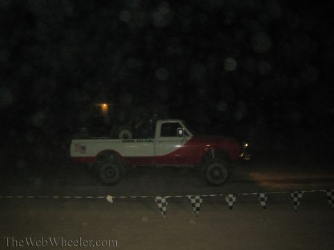 1990 Ford Bronco - two-tone red-and-white lifted pickup with a US flag decal rolls past checkered banner flags in the dark.