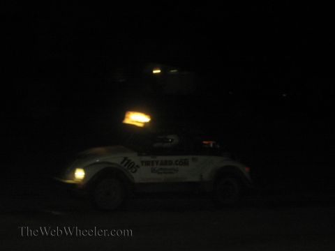 1990 Ford Bronco - white VW Beetle race car numbered 1105 with glowing roof lights speeds through the dark at an off-road event.