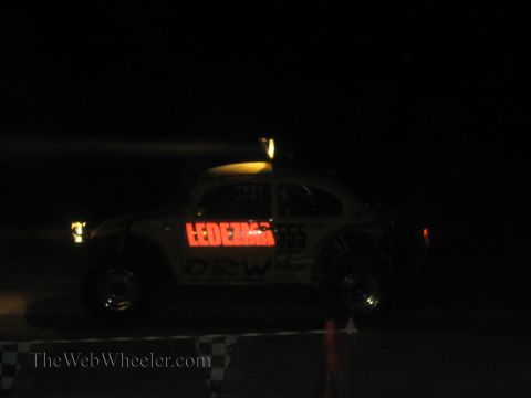 1990 Ford Bronco - white Baja Bug racer marked 'LEDEZMA' and number 55 sits in dim light behind a checkered flag line.