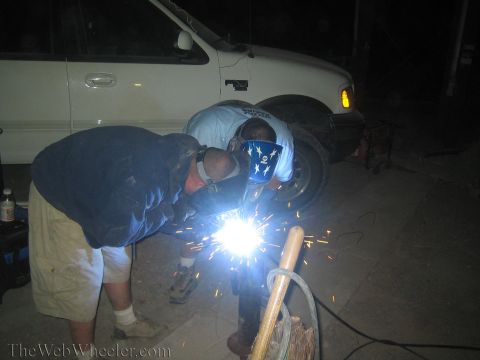 1990 Ford Bronco - Two men in welding masks work on a metal post at night, throwing bright blue sparks beside a white pickup.