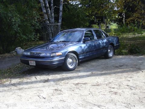 1996 Ford Crown Victoria - A blue sedan sits parked outdoors on a dirt surface, with trees and foliage surrounding it.
