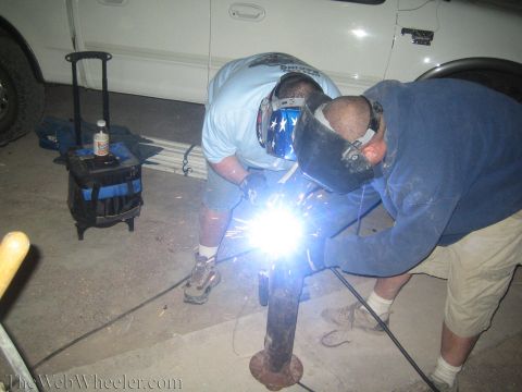 1990 Ford Bronco - Two men in welding masks work close together on a rusty post, sparks flying, beside a white pickup at night.