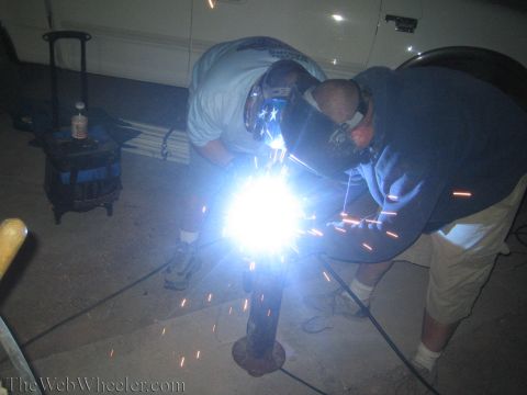 1990 Ford Bronco - Two masked men weld a rusty metal post at night, a bright burst of sparks lighting their hands and the ground.