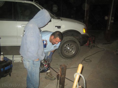 1990 Ford Bronco - man in a gray hoodie bends over a metal post at night beside a white pickup raised on a jack stand.