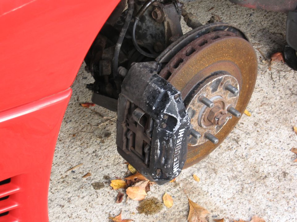 close-up of a car's front brake caliper with 'MITSUBISHI' text, showing a rusty rotor and part of a red fender