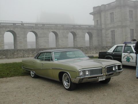 1991 Ford Crown Victoria - A green 1967 Buick LeSabre sedan is parked in front of a stone structure with a highway patrol car behind it.