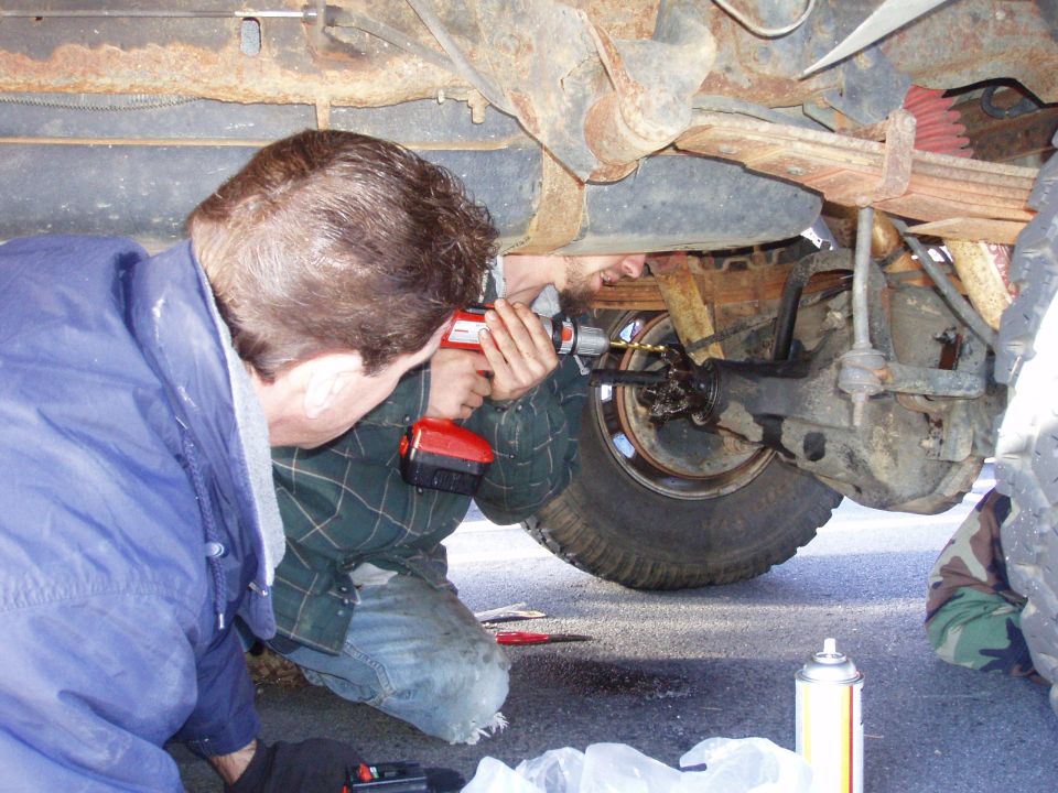 Two men working on the rear axle of a truck, one using a drill