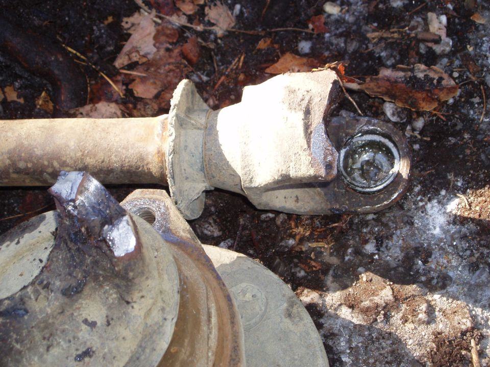 Close-up of a damaged driveshaft U-joint and flange, with dirt and leaves on the ground