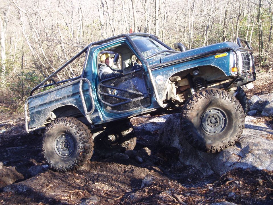 Modified pickup truck with roll cage climbing over rocks off-road. The front grille and body style suggest a late 1970s Ford F-Series