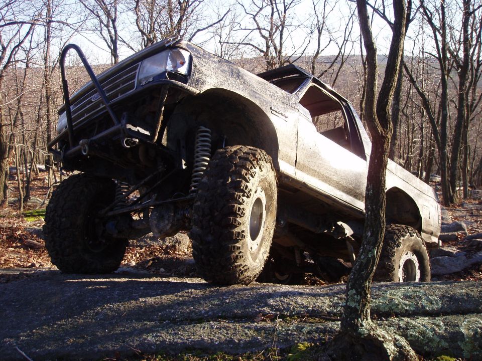 Modified pickup truck with large tires and suspension, driving over rocks in a wooded area