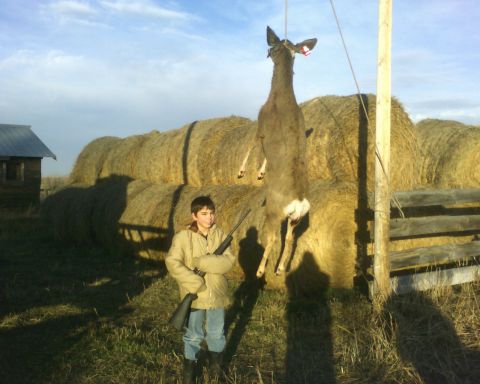 1989 Ford Bronco - A young hunter stands with his rifle next to a deer hanging from a pole, with hay bales and a small building in the background.