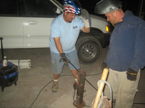 1990 Ford Bronco - man in a US-flag bandana grinds a rusty metal post at night while a welder in a mask watches, by a white truck.