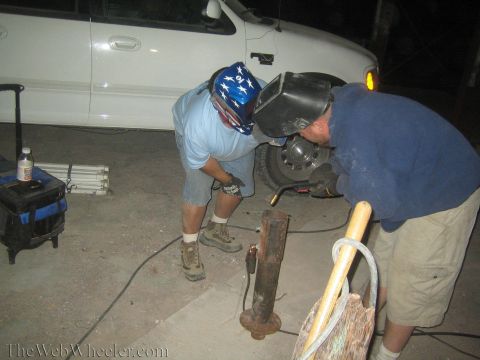 1990 Ford Bronco - man in a stars-and-stripes bandana and a masked welder bend over a rusty metal post in the dark by a white truck.