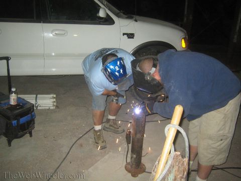 1990 Ford Bronco - Two masked welders work a tall rusty post at night, a violet welding arc and sparks glowing between them.