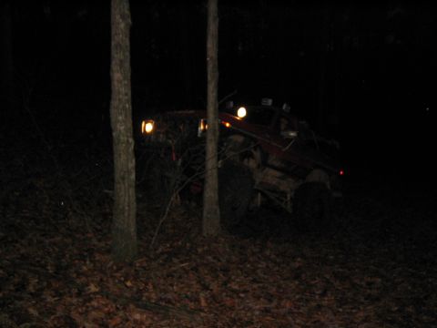 1979 Ford Bronco - A red off-road vehicle navigates a wooded area at night, its auxiliary lights illuminating the path.
