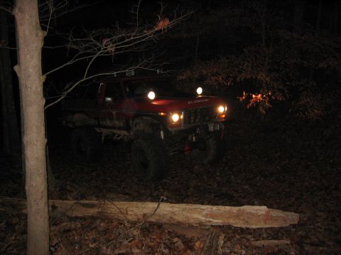 1979 Ford Bronco - A red 1979 Ford Bronco is driven through a wooded area at night, its headlights and auxiliary lights illuminating the path.