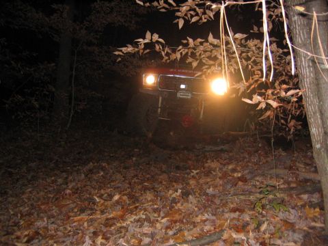 1979 Ford Bronco - A red SUV is parked on a wooded hill at night, its headlights illuminating the surrounding trees and fallen leaves.
