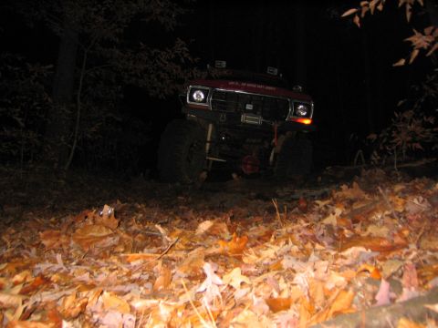 1979 Ford Bronco - A red off-road SUV with large tires and a winch is parked on a leaf-covered ground at night.