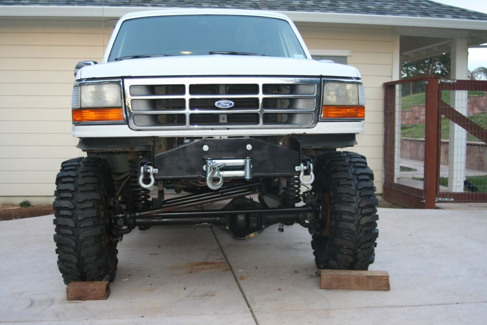 Front view of a lifted white Ford truck with large tires and a winch bumper. The grille has a Ford logo