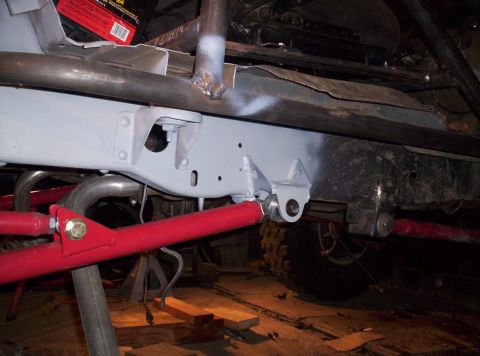 1984 Ford Bronco - Underbody shot of a light-gray painted frame rail with freshly welded gray bracket tabs holding the rod end of a red-painted suspension lin…