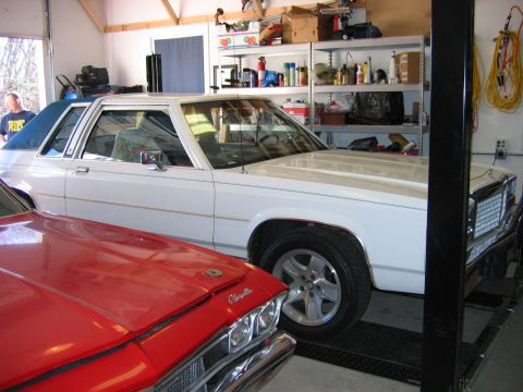 1985 Ford LTD Crown Victoria - A white Ford Crown Victoria is parked on a lift in a garage, with a red car partially visible in front.