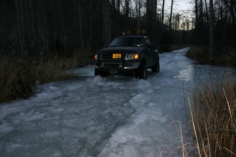 2003 Jeep TJ - A black pickup truck navigates a partially frozen road surrounded by trees.