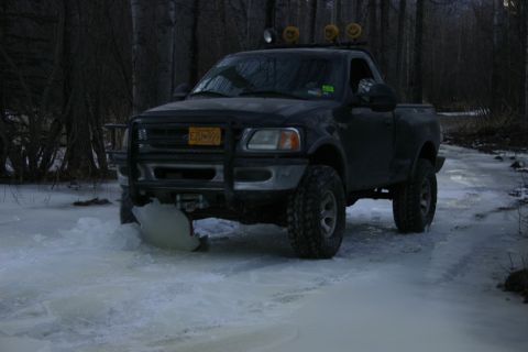 2003 Jeep TJ - A dark-colored pickup truck navigates a slippery, icy path through a forest during daylight.