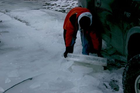 2003 Jeep TJ - A person in an orange jacket works with ice blocks next to a dark truck in a frozen environment.