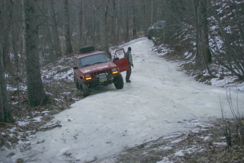 2003 Jeep TJ - A red 2003 Jeep TJ navigates an icy road in a wooded area, with another vehicle visible further up the path.