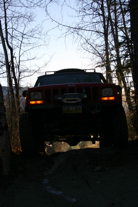 2003 Jeep TJ - A red Jeep Cherokee XJ is seen from the front, driving on a dirt path surrounded by trees.