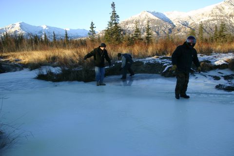 2003 Jeep TJ - Three individuals are seen on a frozen body of water, with a backdrop of trees and snow-covered mountains under a clear blue sky.