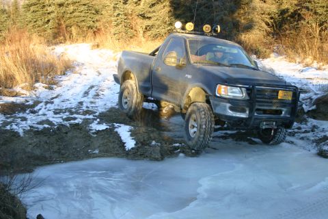 2003 Jeep TJ - A black Ford F-150 pickup truck drives through a muddy and icy terrain with trees in the background.