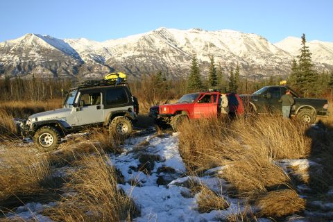 2003 Jeep TJ - A silver 2003 Jeep TJ is parked in a snowy, grassy area with mountains in the background, alongside other vehicles.