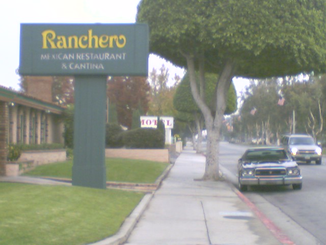 Sign for Ranchero Mexican Restaurant & Cantina next to a sidewalk and street with two cars