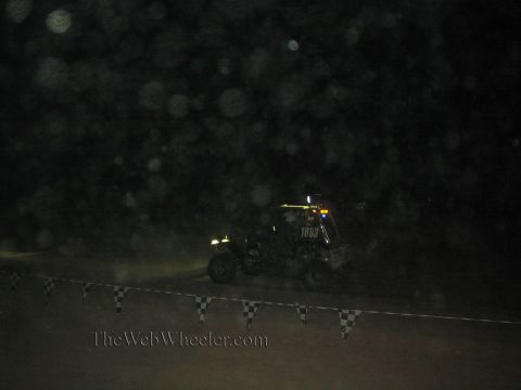 1990 Ford Bronco - off-road race buggy with illuminated roof lights crosses a dark dusty course at night beyond a string of checkered flags.