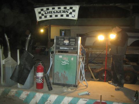 1990 Ford Bronco - At night a person works under bright lamps near an old fuel pump and a red fire extinguisher beneath a Checkers banner.