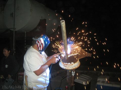 1990 Ford Bronco - person wearing a stars-and-stripes welding hood cuts a vertical metal pipe at night, throwing a bright shower of sparks.