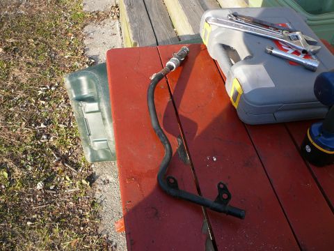 1997 Mercury Grand Marquis - A removed black metal coolant crossover tube with two bolt-flange mounting tabs, a barbed hose nipple and a threaded fitting at the top end…