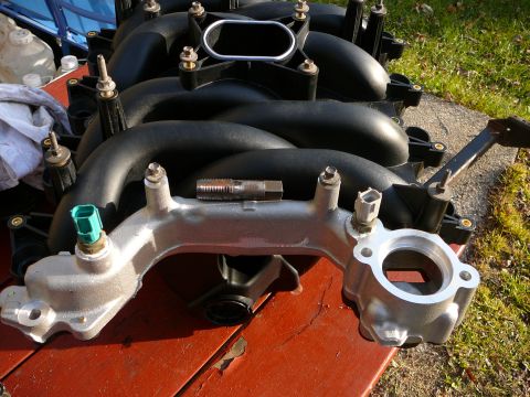 1997 Mercury Grand Marquis - A black plastic upper intake manifold seated on a cast-aluminum lower intake, resting on a red wood bench outdoors. A green two-wire temper…