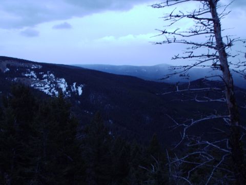 1990 Ford Bronco - A vast mountain landscape is viewed at dusk, with dark evergreen forests and patches of snow on the slopes.