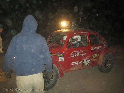 1990 Ford Bronco - hooded person stands beside a muddy red Volkswagen Beetle off-road race car numbered 1146 with roof lights on at night.