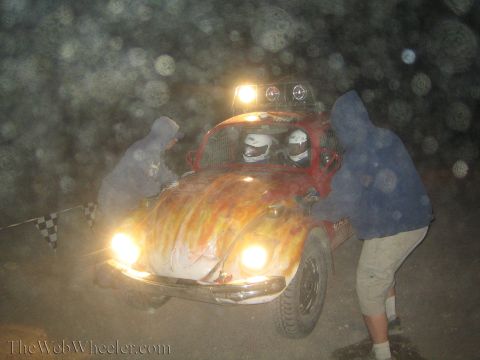 1990 Ford Bronco - Two people work on a flame-painted Volkswagen Beetle off-road race car with headlights and roof lights blazing in the dusty dark.