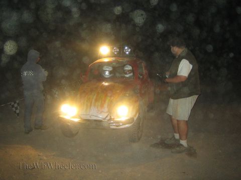 1990 Ford Bronco - flame-painted Volkswagen Beetle off-road racer sits with lights on in a dusty night scene as two people stand nearby.