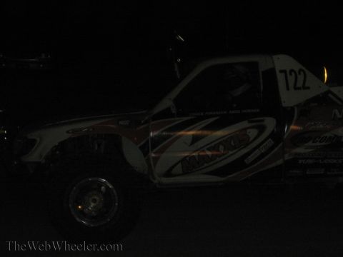 1990 Ford Bronco - white off-road trophy-style race truck numbered 722 with Maxxis livery sits dimly lit in near darkness.