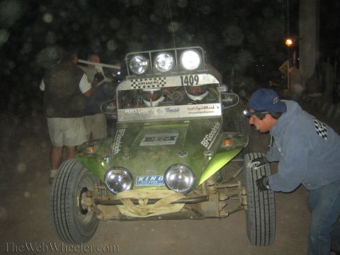 1990 Ford Bronco - People crouch around a green off-road buggy numbered 1409 with its head and roof lights lit during a dusty night stop.