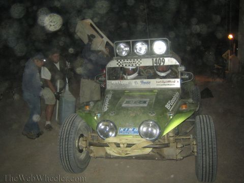 1990 Ford Bronco - crew works around a green off-road buggy numbered 1409, one holding a fuel jug, with lights on during a dusty night pit stop.