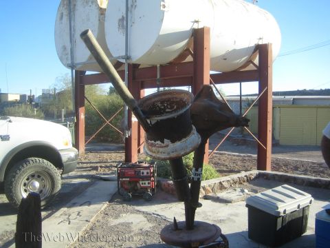 1990 Ford Bronco - improvised welded metal funnel and pipe rig stands under a white tank on a frame, near a portable generator and a white pickup.